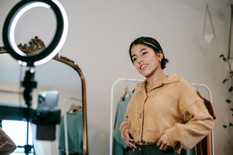 Low angle of positive ethnic female blogger shooting video on cellphone on tripod with shining lamp